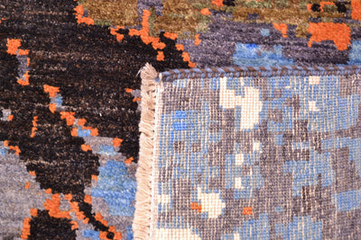 Close-up of the hand-knotted back of a handmade abstract carpet corner showing a cream fringed edge, with splashes of blue, grey, black, and orange woven textile details.