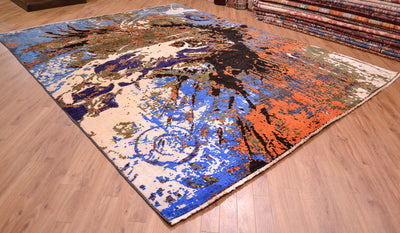 A rectangular modern Abstract area rug with an abstract, distressed artistic splash pattern in blue, orange, beige, black, and brown, displayed on a light wood floor with other rugs stacked in the background.