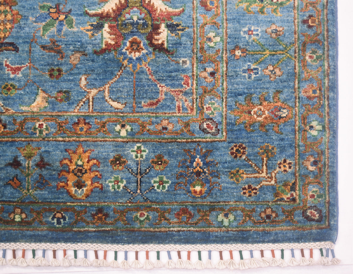 Corner detail of blue handmade rug with floral curvilinear designs with latticed fringes bound colourfully as a finishing features.
