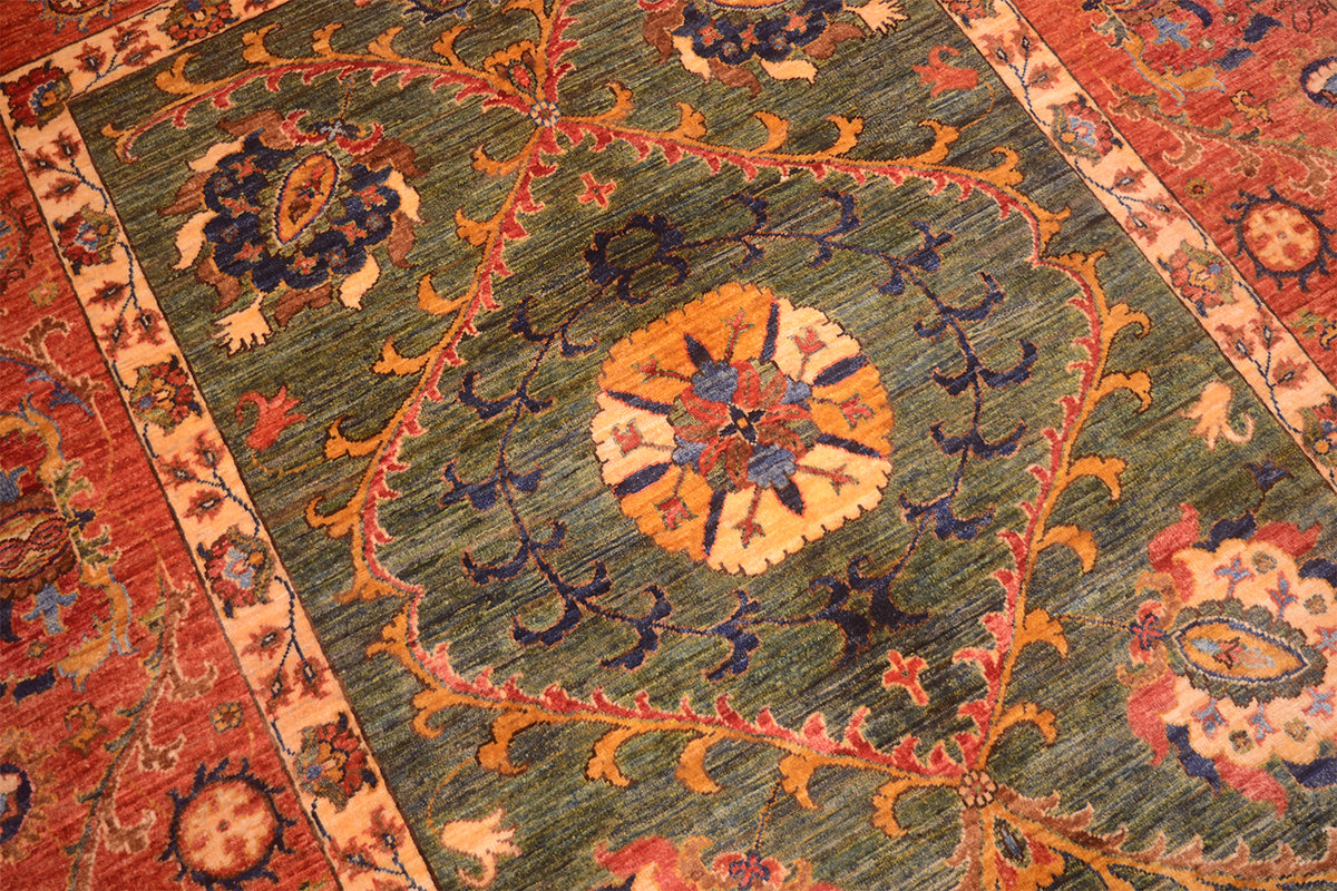 A close-up of an Afghan Suzani design handmade carpet with a muted green field, featuring a central floral medallion in orange, cream, and blue, surrounded by ornate vine motifs and a red border.