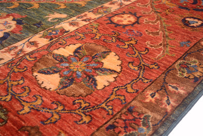 A close-up of an Afghan Suzani design handmade carpet with a warm rust-red field, beige medallion, and blue and green floral details.