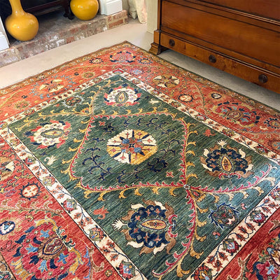 A traditional Afghan Suzani design handmade carpet with a green central field, red borders, and multicoloured floral motifs, shown on a floor in a room with a wooden chest and yellow vases.