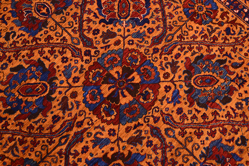 A close-up of a very fine handmade Suzani rug with a warm amber ground and detailed floral medallions in blue, burgundy red, and dark brown.