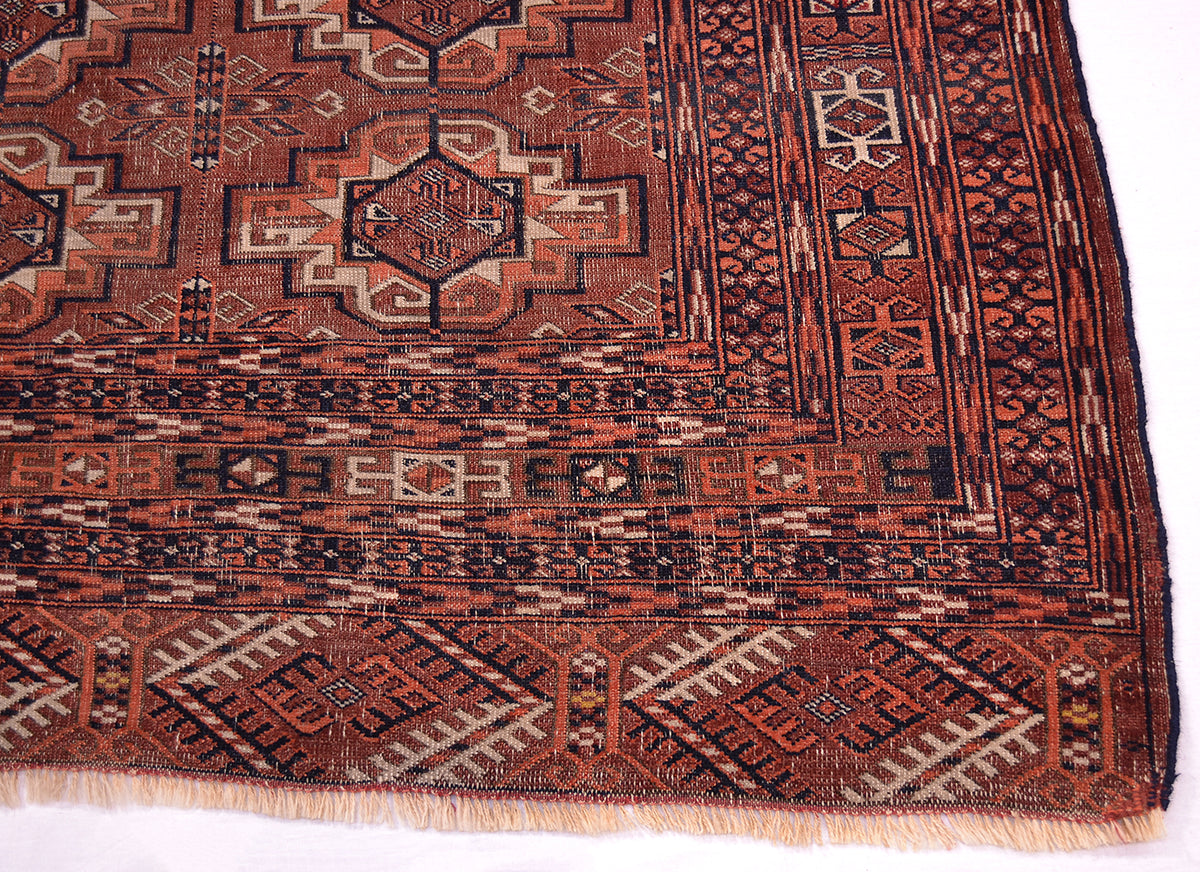 A close-up of a traditional patterned antique Tekke rug corner showing a geometric design in rust red, black, and beige with fringed edges.