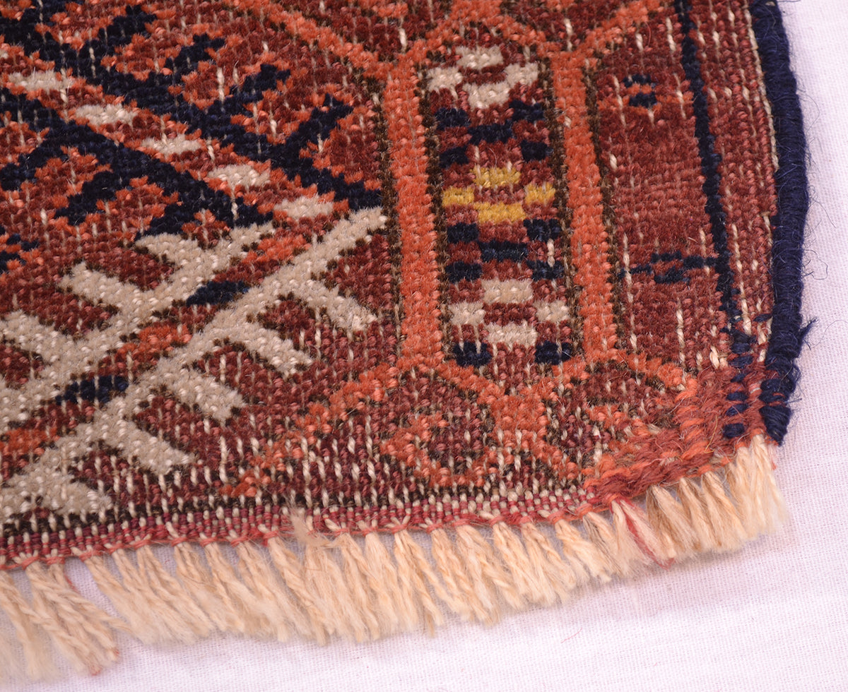 Close-up of a repaired corner of a handwoven antique Tekke rug showing a geometric pattern in rust red, beige, black, and orange with cream fringe.