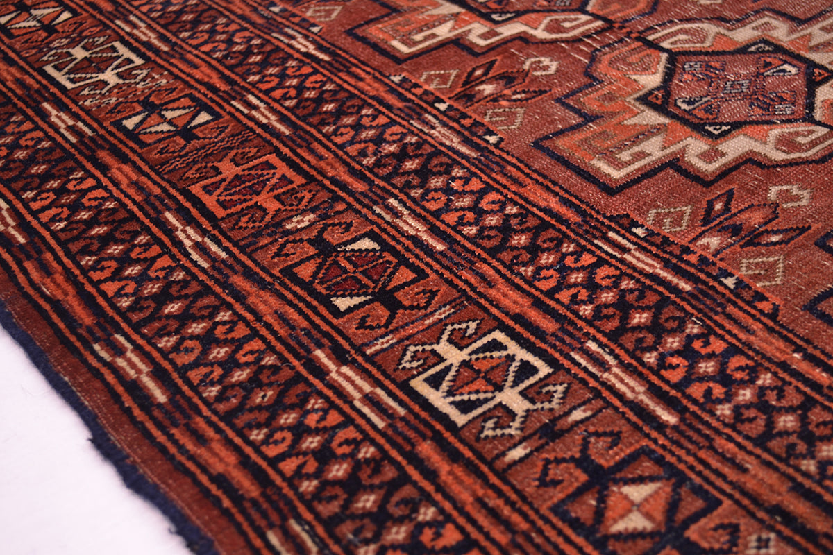 A close-up of a antique Tekke rug with a terracotta red and dark brown base, featuring geometric borders and motifs in beige, black, and rust orange.