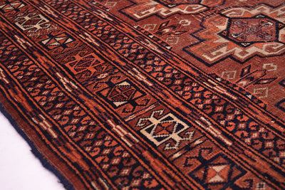 A close-up of a antique Tekke rug with a terracotta red and dark brown base, featuring geometric borders and motifs in beige, black, and rust orange.