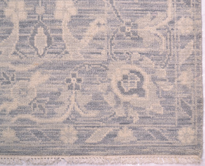 A close-up of a woven tonal rug showing its detailed pattern and texture. The rug features a muted, low-contrast design in dusty blue-gray and warm cream, with a fringed edge visible at the bottom.