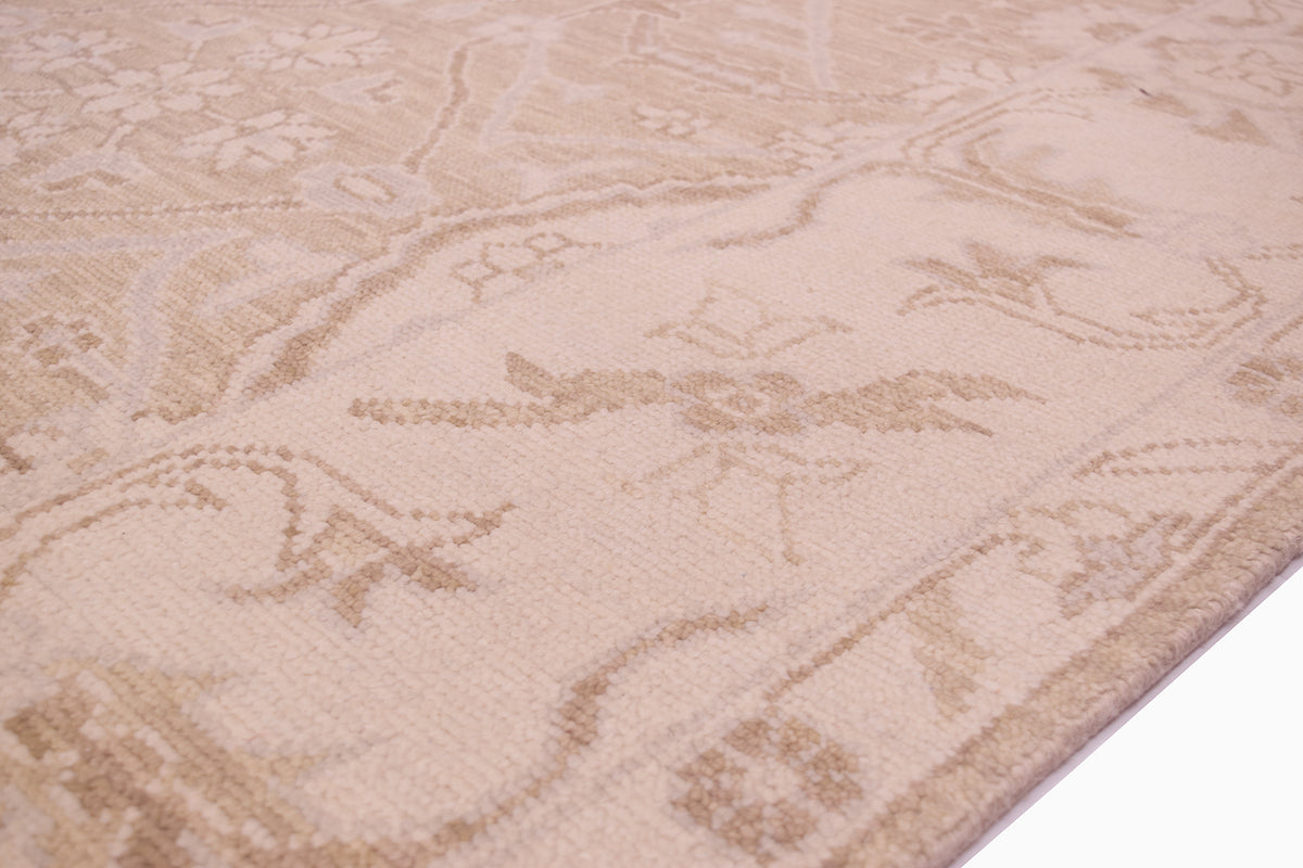A close-up of a beige and cream patterned tonal area rug with a traditional floral and scroll motif, shown with a white surface visible in the bottom right corner.