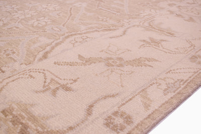 A close-up of a beige and cream patterned tonal area rug with a traditional floral and scroll motif, shown with a white surface visible in the bottom right corner.