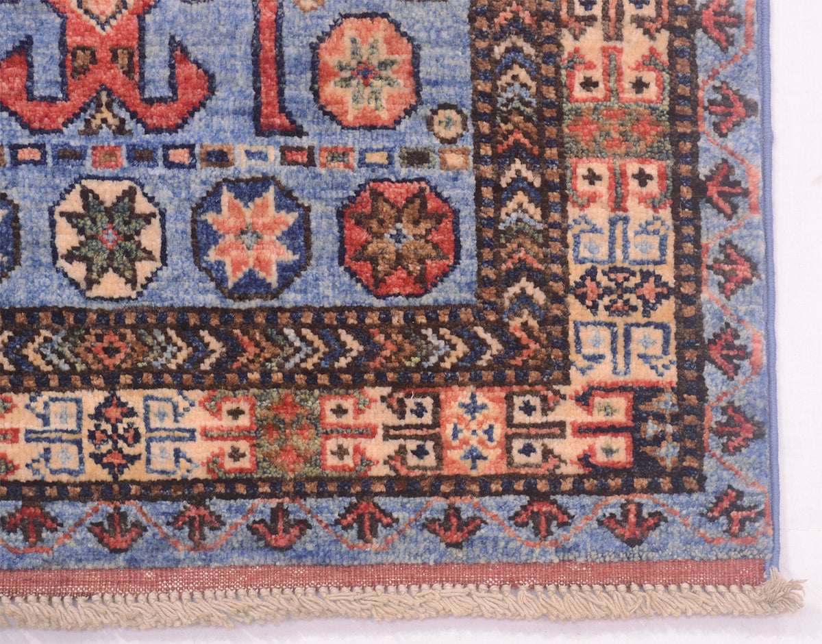 A close-up of a patterned Afghan Turkmen runner corner showing a blue field with geometric and floral motifs in red, beige, and dark brown, plus a fringed edge.