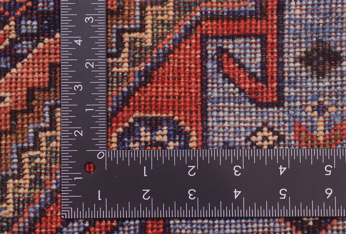 Close-up of the hand-knotted weave of a Turkmen tribal style runner patterned rug with a metal ruler placed along the edge to show scale of the knot density, featuring a geometric design in rust red, blue, beige, and black.