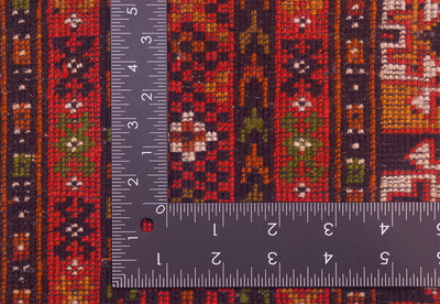 Close-up of a hand-knotted patterned unusual Turkoman Prayer Rug with a metal ruler placed over it to show scale of the knotting. The rug has vertical bands of red, orange, green, black, and cream geometric and floral motifs.