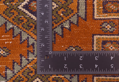 Close-up of a patterned rug mat with rust orange, olive green, beige, and dark accents, shown with a black metal L-shaped ruler to indicate scale.