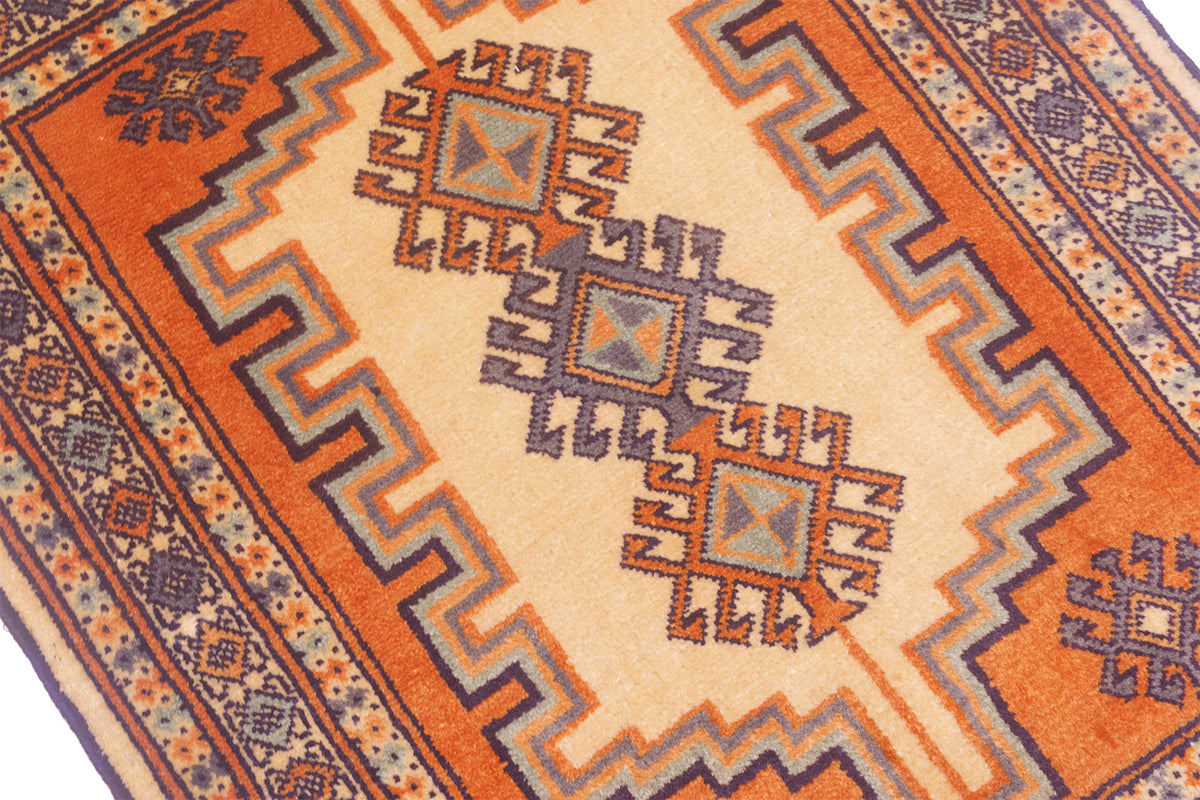 A close-up of a patterned rug with a cream center, orange field, and geometric motifs in blue, brown, and orange.