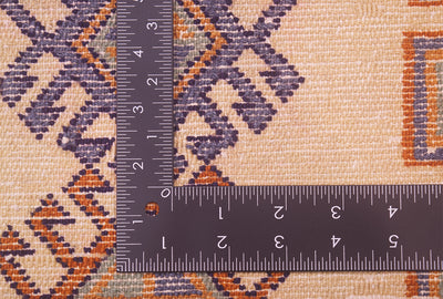 Close-up of a patterned textile with a dark metal L-square ruler placed over it to show scale. The rug mat has a warm beige base with geometric motifs in dark blues and orange.