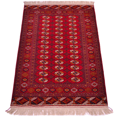 A traditional oriental Turkoman Bokhara rug with a deep red field, featuring a central row of repeating Tekke gul motifs in cream, orange, and black, surrounded by ornate borders and cream fringe on the top and bottom.