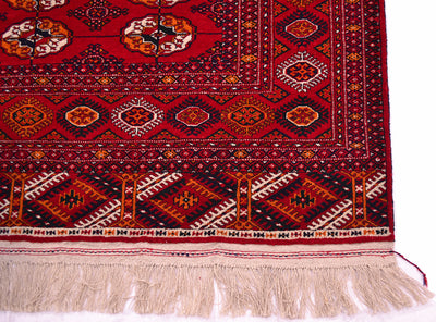 A close-up of a patterned Bokhara Turkoman rug corner showing a deep red field with geometric and floral motifs in black, beige, orange, and white, finished with a cream-coloured fringe.