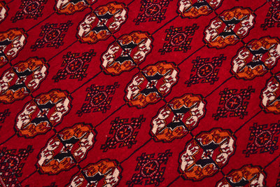A close-up of a Turkoman Bokhara red rug featuring repeating ornate tribal gul motifs in orange, cream, and black.
