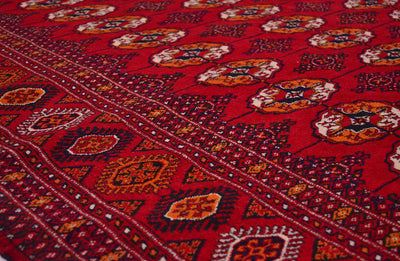 A close-up of a red patterned Bokhara Turkoman rug with geometric borders and floral motifs in orange, cream, and dark blue.