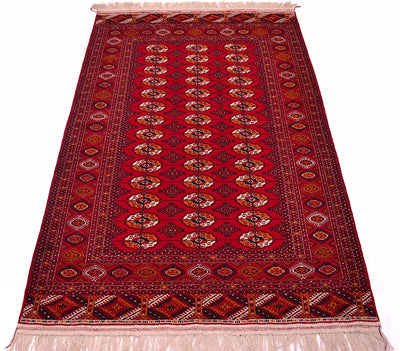 A traditional red Turkoman Bokhara wool rug with a central field of repeating Tekke gul motifs, bordered by ornate geometric and floral patterns, and finished with cream-coloured fringe on both ends.