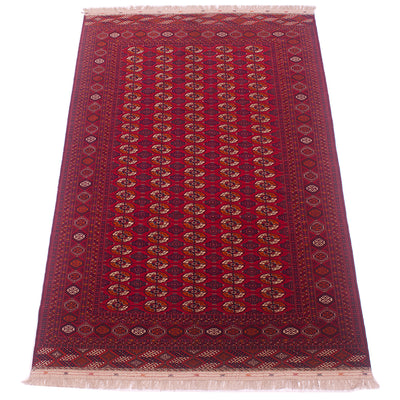 A traditional rectangular Turkoman Bokhara Carpet with a deep red field, featuring vertical columns of repeating medallion motifs in beige, brown, and blue, surrounded by a detailed patterned border and cream fringe on the ends.