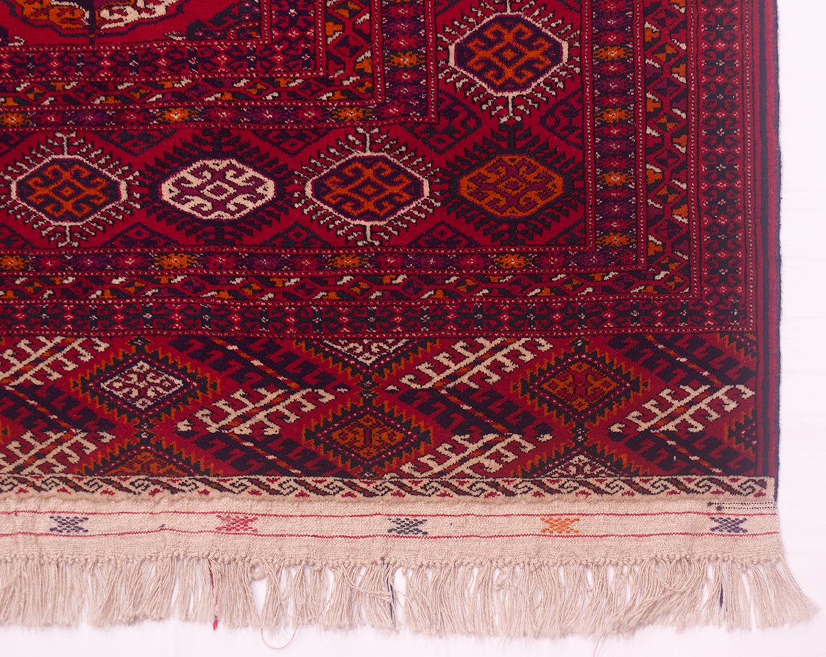 A close-up of a traditional patterned Turkoman Bokhara Carpet with a deep red field, geometric diamond motifs in orange, beige, and black, and a fringed cream-coloured flatweave end at the bottom.