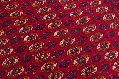 A close-up of a red patterned Bokhara Turkoman carpet with repeating geometric and floral motifs in orange, beige, and dark purple.