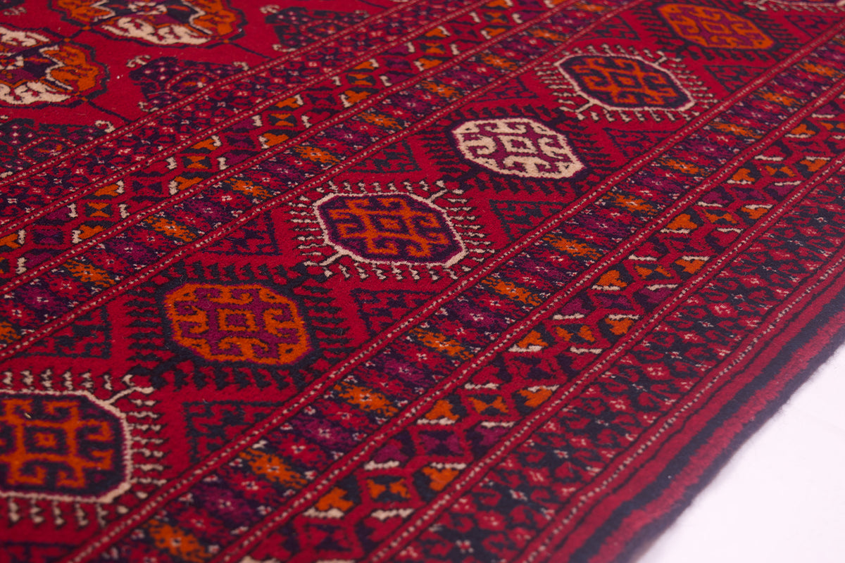 A close-up of a traditional patterned Turkoman Tekke Bokhara carpet with a deep red field, geometric borders, and repeating medallion motifs in orange, cream, and dark blue.