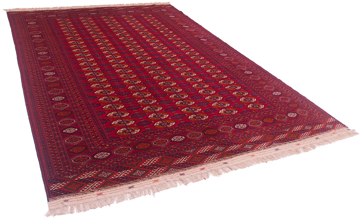 A traditional Bokhara-style Turkoman wool carpet with a deep red field, intricate geometric and floral patterns in navy, beige, and gold, and cream-coloured fringe on the ends.