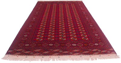A traditional style Turkoman Bokhara wool rug with a deep red field, intricate geometric and floral motifs in navy, beige, and brown, and cream fringe on the ends.