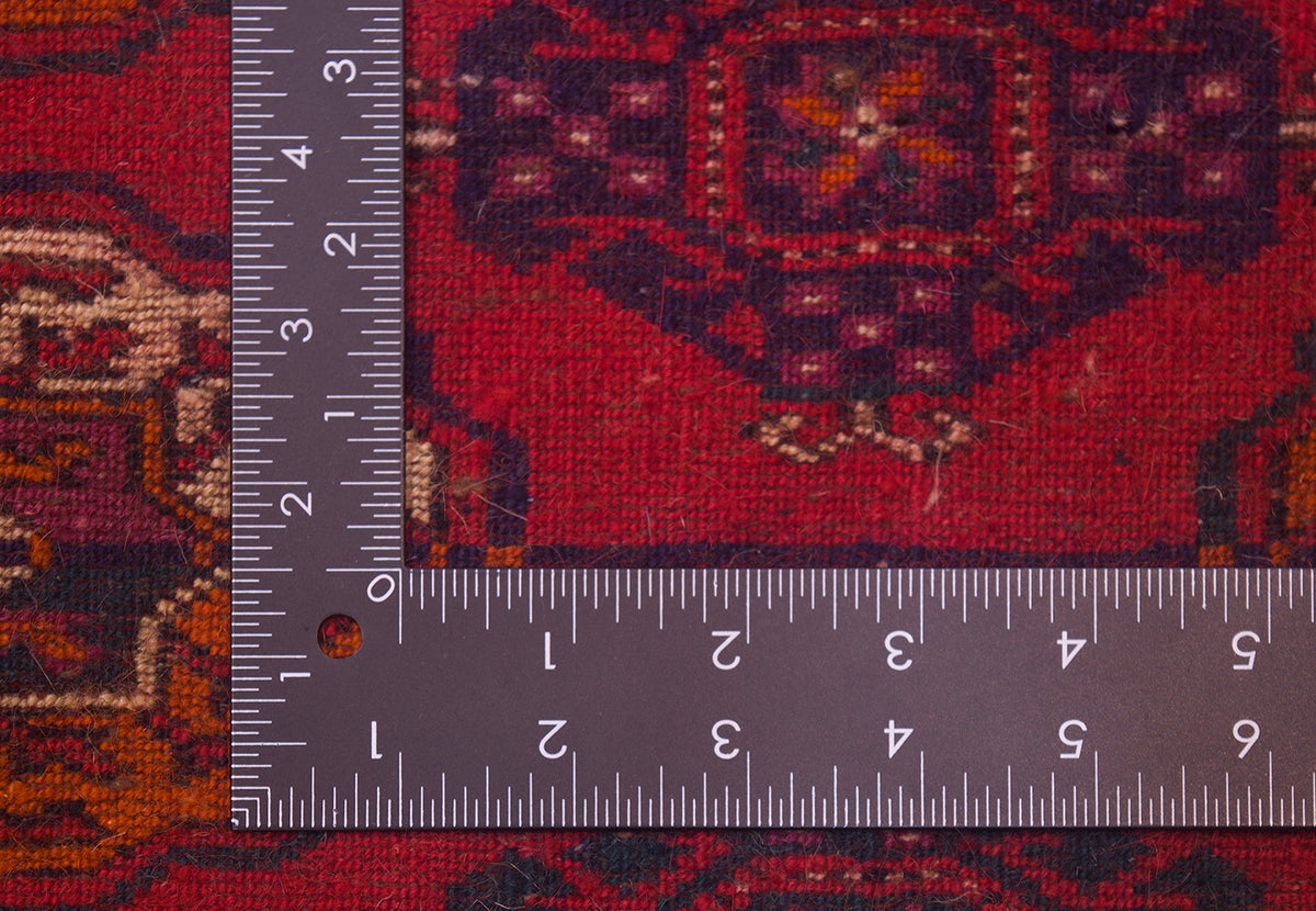A close-up of the hand-knotted weave of a red Turkoman Bokhara Carpet with a dark L-shaped metal ruler placed along the corner to show the scale of the knotting.