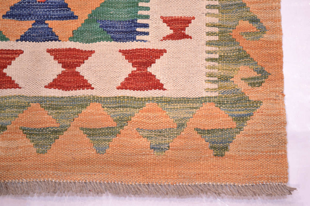 Close-up of a handwoven Kilim runner rug corner showing a cream base with geometric patterns in terracotta red, muted green, and blue, finished with a fringed edge.