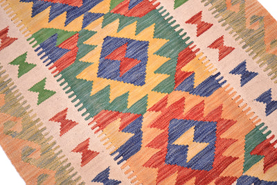A close-up of a woven kilim rug featuring a vibrant geometric pattern with orange, blue, green, and red shapes on a beige background.