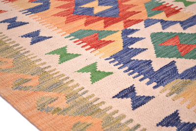 A close-up of a woven kilim runner rug showing a geometric pattern with blue, green, red, and yellow motifs on a warm beige background.