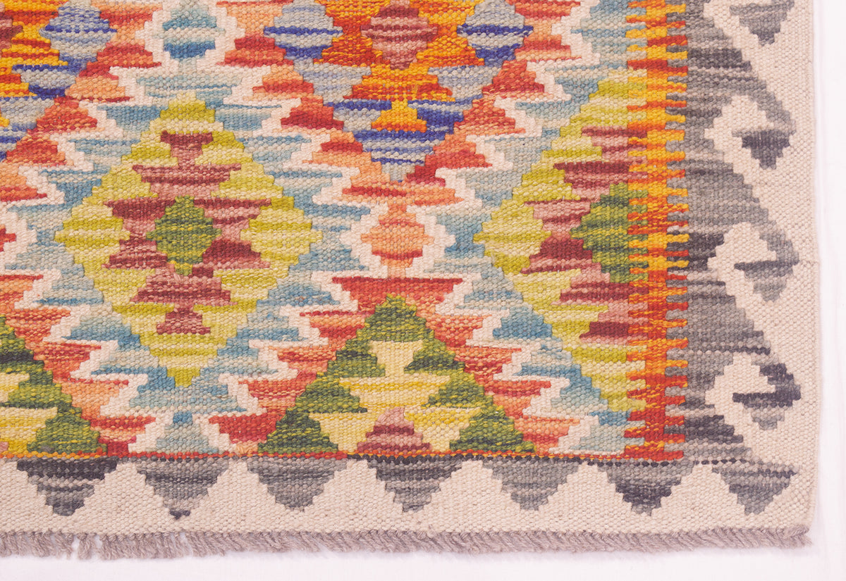 Close-up of a handwoven Afghan kelim rug corner showing a multicolour geometric pattern with diamonds and zigzags in yellow, green, orange, blue, red, cream and grey on a white base.