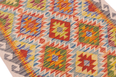 A close-up of a handwoven Afghan Kelim rug with a colourful geometric pattern, featuring repeating diamond motifs in red, yellow, green, blue, orange, beige and grey on a white background.