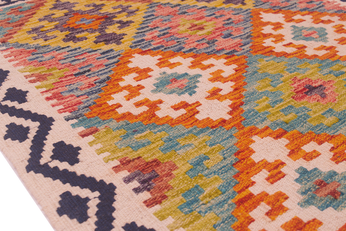 A close-up of a handwoven Kilim rug with a colourful geometric pattern in orange, pink, blue, yellow, and cream, featuring a dark navy patterned border on a white background.