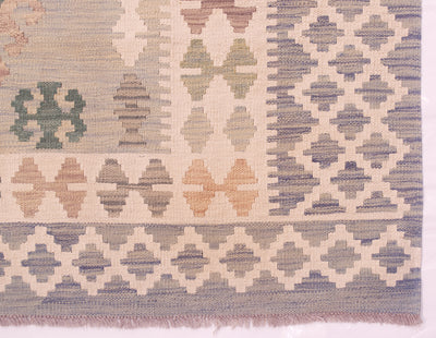 A close-up of a handwoven Afghan Veg Dye Kilim rug with a geometric design in muted blue-grey, beige, cream, and soft green tones, finished with a fringed edge.