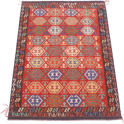A flat-laid kilim rug with a vibrant red field, decorated with repeating geometric diamond and hexagon motifs in blue, beige, and orange, framed by a dark patterned border.