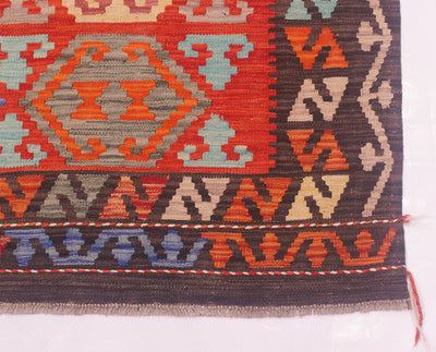 A close-up of a Red Afghan Kilim Rug corner showing geometric patterns in red, orange, brown, beige, and light blue on a white background.