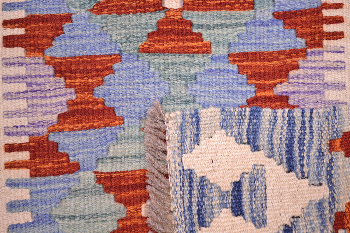 Close-up of the weave from a handwoven Afghan Kilim runner rug with a geometric pattern in blue, rust orange, and light blue, featuring a folded corner showing a cream and blue striped section with grey fringe.