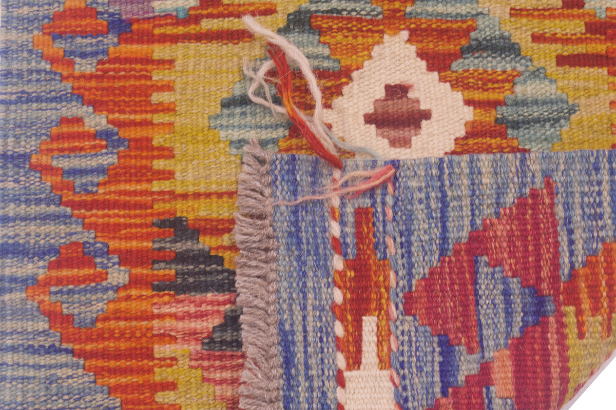 Close-up of the weave of a handwoven Afghan Kilim strip with a geometric pattern in orange, blue, yellow, red, and beige, showing a frayed edge with loose threads.