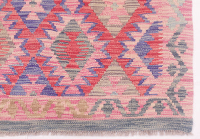 A close-up of a woven kilim Afghan rug showing a geometric diamond pattern in pink, coral, purple, and grey, with a fringed edge at the bottom.