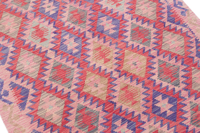 A close-up of a geometric kilim-style patterned rug in pink, red, blue, beige, and grey, shown on a white background.