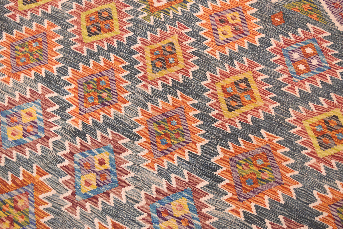 A close-up of a handwoven Afghan Large Kilim with a repeating geometric kilim-style pattern in orange, yellow, blue, green, and cream on a dark green background.