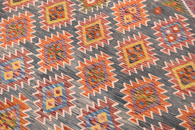 A close-up of a handwoven Afghan Large Kilim with a repeating geometric kilim-style pattern in orange, yellow, blue, green, and cream on a dark green background.