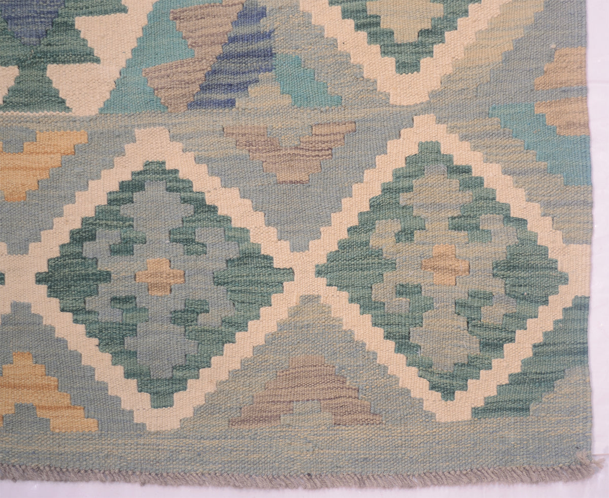 A close-up of a handwoven Kilim rug with a geometric diamond pattern in muted teal, sage green, beige, and light blue tones, showing the textured weave and fringe along the bottom edge.