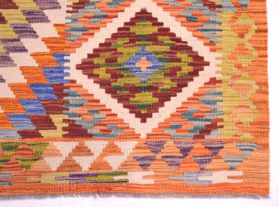 A close-up of a woven Afghan kilim rug showing a geometric diamond pattern in orange, brown, green, blue, and cream.