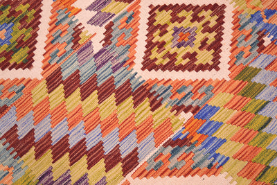 Close-up of a woven Afghan Kilim rug with a geometric pattern in orange, brown, yellow, blue, green, and purple on a light pink ground.